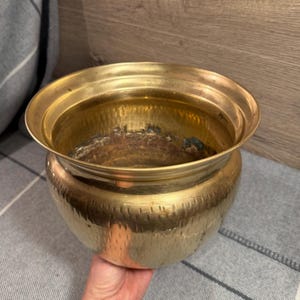May include: A round brass planter with a wide, flared rim and a textured, hammered exterior finish. The interior of the vessel shows signs of oxidation and dark patina, contrasting with the polished, reflective golden surface of the metal body.