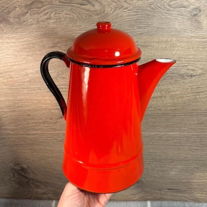 Könnte beinhalten: Eine leuchtend rote Emaille-Kaffeekanne mit glänzender Oberfläche, schwarzem Griff und passendem rotem Deckel mit kleinem Knauf. Das Gefäß hat einen konisch zulaufenden zylindrischen Körper, einen breiten Boden und einen markanten Ausguss mit weißem Rand.