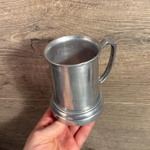 May include: A silver-colored metal mug with a handle. The mug has a cylindrical body and a slightly flared base. The handle is rectangular and attached to the side of the mug. The mug is held in a person's hand.