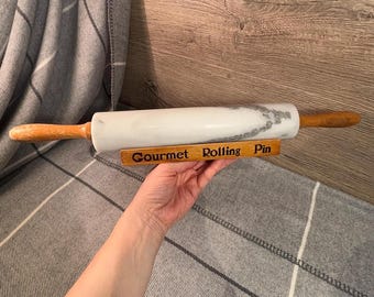 May include: White marble rolling pin with a wooden handle. The rolling pin is resting on a wooden stand with the words "Gourmet Rolling Pin" engraved on it.