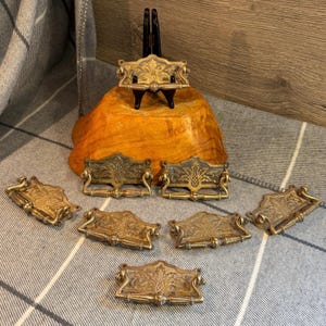 May include: A set of antique brass drawer pulls featuring intricate floral and leaf patterns. The pulls, in various sizes, have a warm, aged patina. One is displayed on a black stand, the others are arranged on a striped fabric. Measurements are in centimeters.