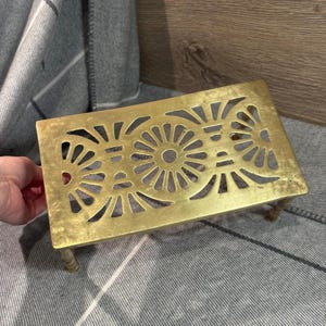 May include: A brass trivet with a decorative design of two large circles with radiating lines. The trivet has four legs and is made of solid brass.