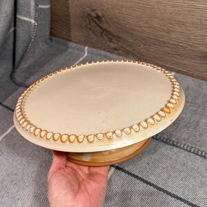 Puede incluir: Un soporte para pasteles de cerámica con una parte superior de color crema y un borde marrón con cuentas. La base es de color marrón a juego. El soporte está diseñado para sostener pasteles y otros productos horneados. El soporte está hecho a mano y tiene una estética rústica.
