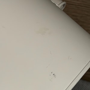 May include: A smooth, off-white curved surface featuring a small, light brown smudge near the center and a cluster of faint, dark blue marks toward the lower edge. A small, white plastic component is visible at the top edge of the object.