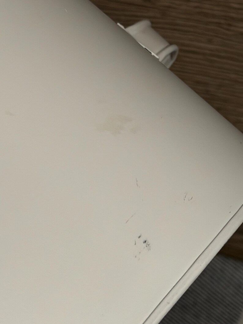 May include: A smooth, off-white curved surface featuring a small, light brown smudge near the center and a cluster of faint, dark blue marks toward the lower edge. A small, white plastic component is visible at the top edge of the object.