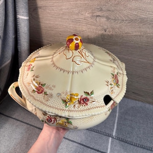 May include: A vintage cream-coloured ceramic serving bowl with a floral design and a lid. The lid has a small knob on top and a small opening on the side. The bowl has a scalloped edge and a floral design.