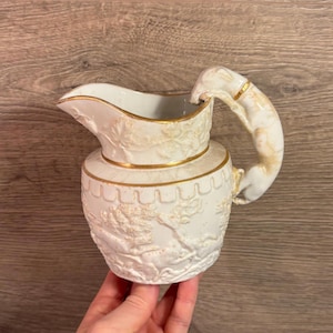 May include: A white ceramic pitcher with a detailed floral relief design and gold trim. The handle is curved and also features gold accents. The pitcher is held by a person's hand.