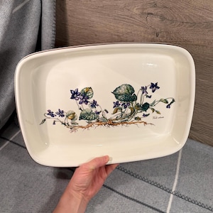 May include: A rectangular off-white ceramic serving platter featuring a botanical illustration of purple violet flowers with green leaves and brown roots. The rim of the platter is outlined with a thin brown line, and the text Viola odorata is printed.