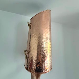 Op de afbeelding: Een koperkleurige, gehamerde metalen emmer met een gebogen bovenkant en twee messing handvatten. De emmer heeft een cilindrische vorm en een getextureerd oppervlak. De handvatten zijn hartvormig. De emmer wordt waarschijnlijk gebruikt voor decoratieve of functionele doeleinden.