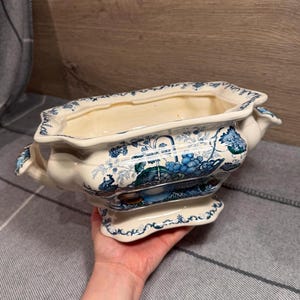 May include: Cream-coloured, rectangular ceramic planter with blue floral and fruit patterns. The planter has a scalloped rim, two handles, and a pedestal base. The design includes images of grapes and foliage.