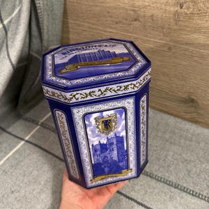 以下が含まれることがあります： 青と金の八角形の紅茶缶。缶には建物のイラストと「RINGTONS LTD TEA MERCHANTS」の文字が描かれています。デザインには、金色の花のアクセントと紋章が含まれています。