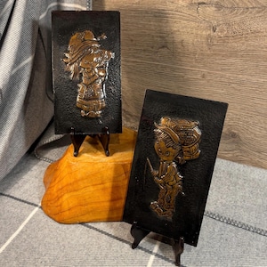 May include: Two vintage copper wall plaques with black backgrounds. Each plaque features a raised relief image of a child. One plaque depicts a girl with a red dress and a white hat. The other plaque depicts a boy with a blue shirt and a brown hat.
