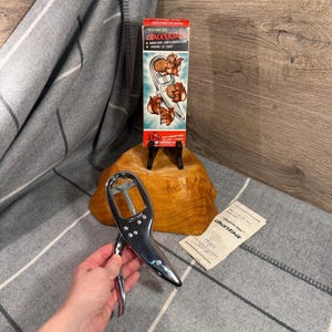 May include: A vintage Cracker Jack box with a red and white design, featuring illustrations of squirrels cracking nuts. A chrome nutcracker is held in a hand, and a paper with text is next to it. The items are on a gray and white striped blanket.