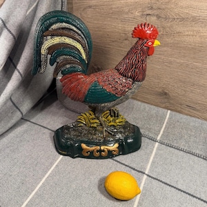 Peut inclure: Une figurine de coq colorée avec une crête rouge, un corps rouge et marron, et des plumes de queue vertes. Le coq est posé sur une base verte avec des détails dorés. Un citron jaune est au premier plan.