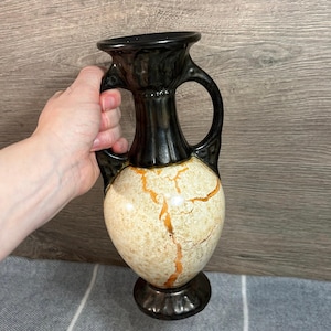 May include: A decorative ceramic vase with a black top and base, and a cream-colored body with orange crackle-like accents. The vase has two handles and a pedestal base. The vase is held by a hand.