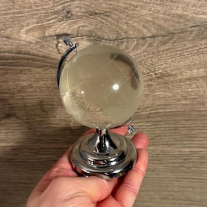 Puede incluir: Un globo terráqueo de cristal transparente con líneas grabadas que representan continentes y países. El globo está montado sobre una base de metal plateado brillante con un soporte curvo. El globo se sostiene en una mano.