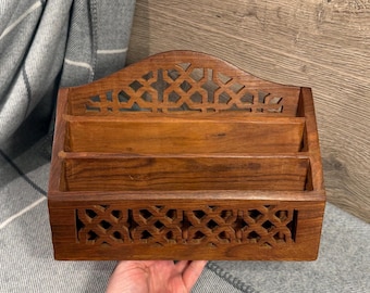 May include: A wooden desk organizer with three compartments. The organizer is made of a dark brown wood and features a decorative geometric cutout design on the front and top. The organizer is held by a person's hand.