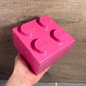 Op de afbeelding: Een felroze LEGO opbergsteen met vier ronde noppen aan de bovenkant, elk met het LEGO-logo. De container wordt in een hand gehouden en is ontworpen om op een klassieke LEGO-steen te lijken.