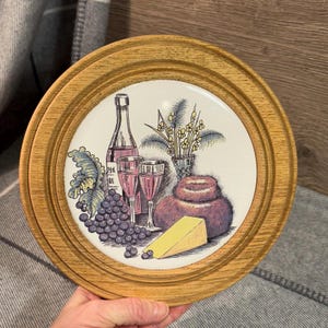 May include: A wooden framed plate with a white ceramic insert featuring a still life of a bottle of wine, two wine glasses, grapes, a wedge of cheese, and a round loaf of bread. The plate has a brown wooden frame with a raised edge.