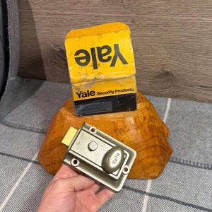 May include: A silver-toned metal Yale rim lock with a thumb turn knob, positioned in front of its original yellow and black cardboard box labelled Yale Security Products. The rectangular lock features four mounting holes and a protruding latch bolt.