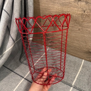 May include: A red wire basket with a heart-shaped design around the rim. The basket is cylindrical and made of thin, red metal wires. The basket is held in a person's hand, with a gray and white plaid blanket in the background.