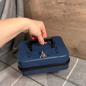May include: A blue metal cash box with a black handle and a key lock. The cash box is sitting on a grey and white plaid blanket. The cash box is approximately 15 cm wide and 20 cm long.