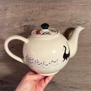 Puede incluir: Tetera de cerámica color crema con una ilustración de un gato negro y un grupo de gatitos esbozados. La tapa presenta un pomo verde y diseños coloridos con temática navideña. La tetera tiene un asa curva y un pico.