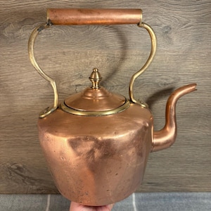 以下が含まれることがあります： 真鍮の取っ手と注ぎ口が付いたビンテージの銅製のやかん。やかんはくすんだ銅の仕上げで、真鍮の蓋にフィニアルが付いています。