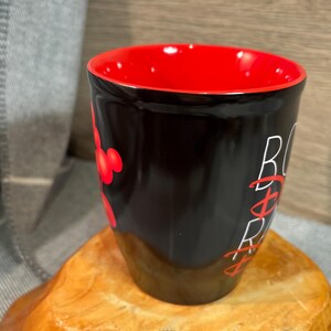 Peut inclure: Une tasse en c&eacute;ramique noire avec un int&eacute;rieur rouge. La tasse pr&eacute;sente le texte "B&Oslash;SS" en blanc et des graphiques rouges de style Mickey Mouse. La tasse est pos&eacute;e sur une surface en bois.