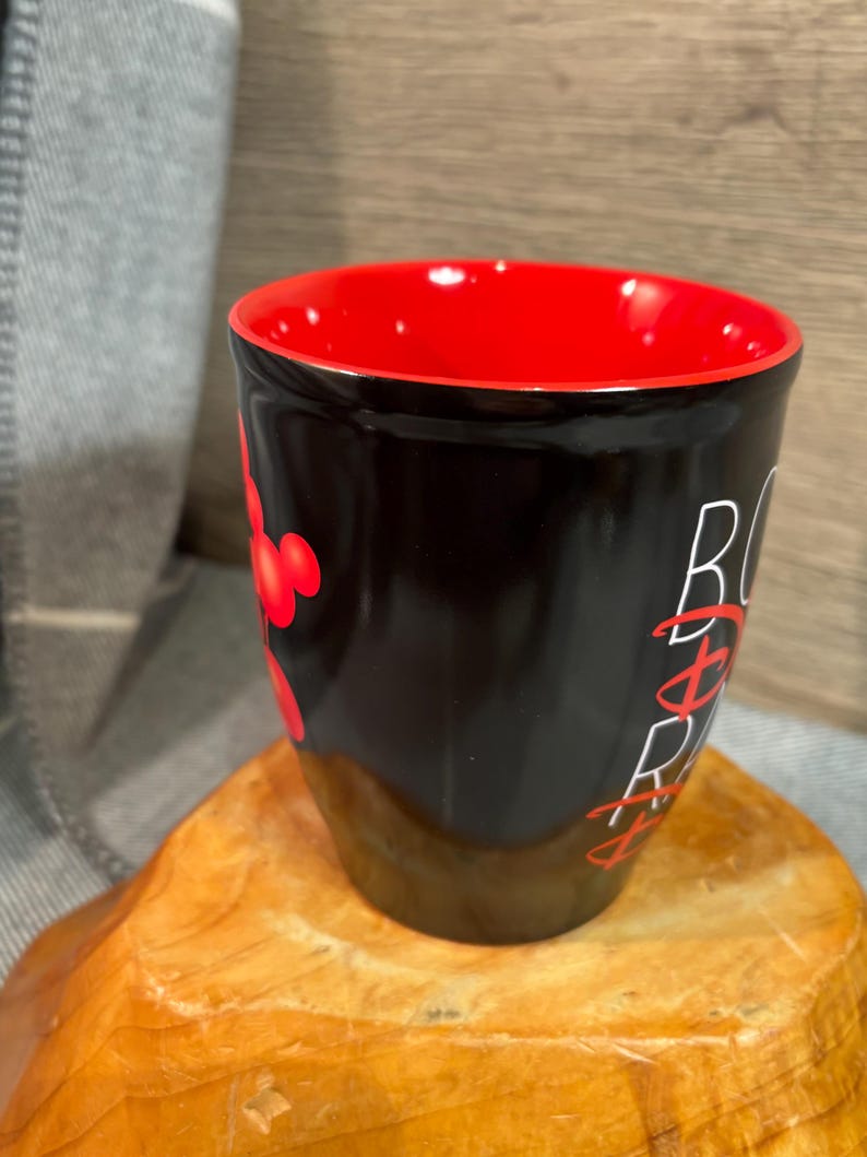 Peut inclure: Une tasse en c&eacute;ramique noire avec un int&eacute;rieur rouge. La tasse pr&eacute;sente le texte "B&Oslash;SS" en blanc et des graphiques rouges de style Mickey Mouse. La tasse est pos&eacute;e sur une surface en bois.