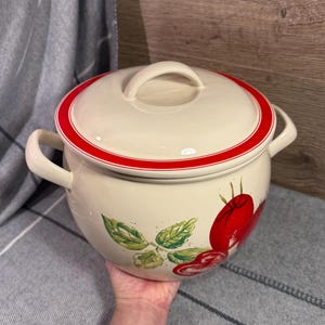 以下が含まれることがあります： クリーム色のセラミック製の調理鍋で、蓋と2つのハンドルが付いています。鍋は赤い縁があり、赤いトマトと緑の葉で飾られています。クラシックなキッチン用品です。