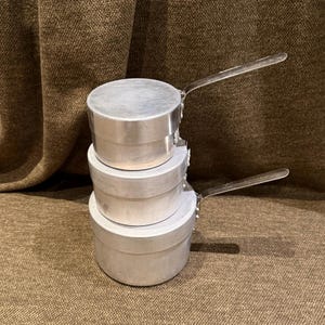 Könnte beinhalten: Ein Stapel aus drei runden, silberfarbenen Aluminiumtöpfen mit Deckeln. Jeder Topf verfügt über einen flachen, geraden Metallgriff an der Seite. Die Töpfe sind in ihrer Größe abgestuft, wobei der kleinste Topf ganz oben steht.