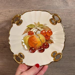 Puede incluir: Un plato de servir de cerámica blanca con ribetes dorados y un diseño pintado de fruta. El plato tiene dos asas a cada lado. El diseño de la fruta presenta melocotones, cerezas y hojas.