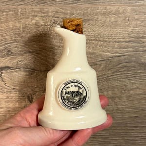 Puede incluir: Una botella de cerámica blanca para aceite y vinagre con un tapón de corcho. La botella tiene una etiqueta en blanco y negro que dice "The original Mason Cash English Kitchenware Company Church Gresley, England  DE11 9QL".