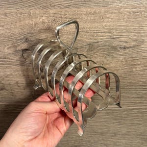 Puede incluir: Un tostador de color plateado con asa. El tostador tiene una serie de ranuras curvas para sostener las rebanadas de pan tostado en posición vertical. El diseño es simple y funcional, con una estética clásica. El tostador es de metal y parece antiguo.