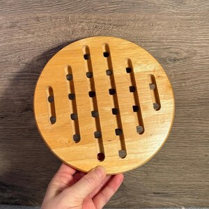 Puede incluir: Un salvamanteles redondo de madera clara con una serie de ranuras verticales y pequeños agujeros espaciados. El salvamanteles está diseñado para proteger las superficies de los platos calientes, sobre un fondo de madera.