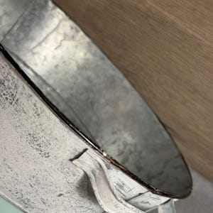 Op de afbeelding: Een ronde emmer van gegalvaniseerd metaal met een verweerde witte verfafwerking aan de buitenkant en een gladde metalen binnenkant. De emmer is voorzien van twee handgrepen aan de zijkant en een gladde, licht mintgroene basis bij de onderrand.