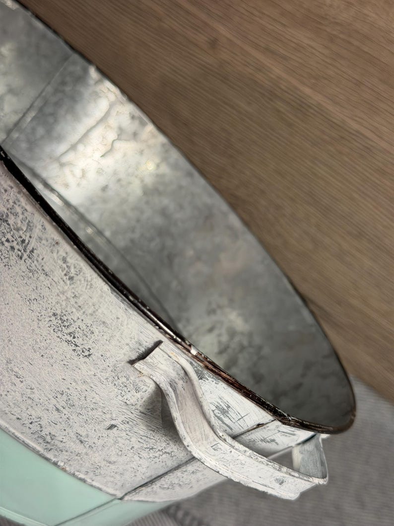 Op de afbeelding: Een ronde emmer van gegalvaniseerd metaal met een verweerde witte verfafwerking aan de buitenkant en een gladde metalen binnenkant. De emmer is voorzien van twee handgrepen aan de zijkant en een gladde, licht mintgroene basis bij de onderrand.