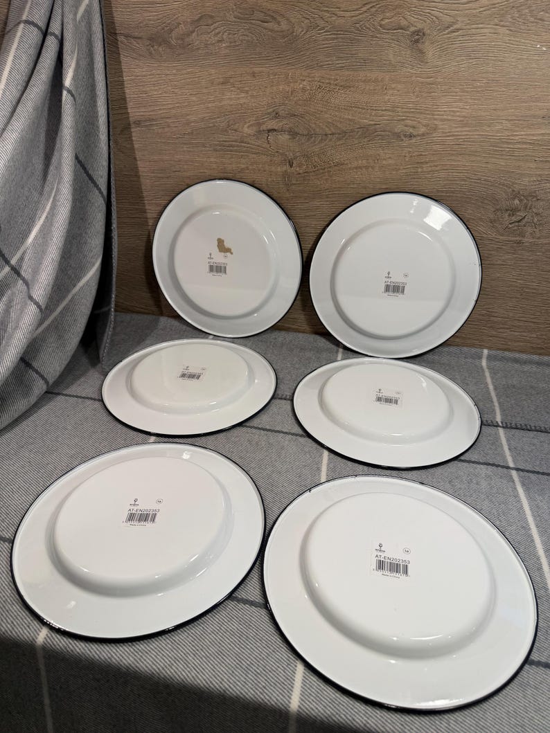 Pode incluir: Um conjunto de seis pratos de esmalte branco com bordas pretas, dispostos em duas filas. Cada prato apresenta uma base circular central, uma borda plana e uma pequena etiqueta retangular com c&oacute;digo de barras na parte inferior.