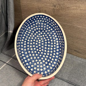 Puede incluir: Cuenco ovalado de cerámica con un patrón de lunares azules y blancos. El cuenco tiene un borde color crema y un interior profundo. El cuenco está sostenido por la mano de una persona.