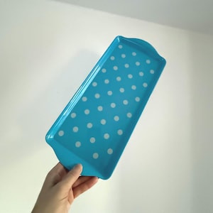 May include: A turquoise serving tray with white polka dots. The rectangular tray has rounded edges and a handle on one end. Ideal for serving snacks or drinks, it measures approximately 30 cm in length.