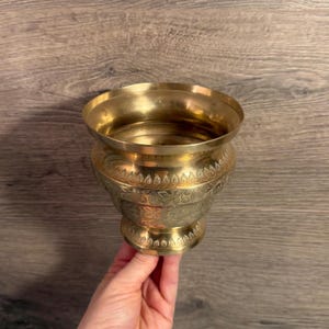 May include: A gold-coloured metal bowl with detailed floral patterns etched around the body. The bowl has a wide, open top and a rounded base. The bowl is held by a hand, and the background is a wooden surface.