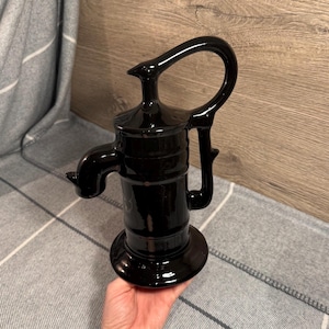 Könnte beinhalten: Eine glänzend schwarze dekorative Wasserpumpe mit gebogenem Griff und Ausguss. Die Pumpe hat einen runden Sockel und eine detaillierte Oberseite. Das Objekt besteht aus einem glänzenden Material.