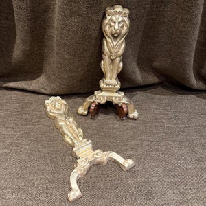 Puede incluir: Un par de morillos de chimenea de metal en tono dorado, cada uno con una figura de león esculpida en la parte superior del poste vertical. La base de cada morillo tiene un diseño curvo y ornamentado con pies en forma de garra.