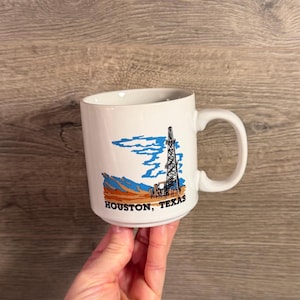 Può includere: Una tazza in ceramica bianca con un'illustrazione grafica di una torre di trivellazione petrolifera nei toni del blu e dell'arancione, su un paesaggio stilizzato. Il testo HOUSTON, TEXAS è stampato in grassetto nero alla base del disegno.
