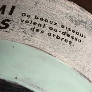 Op de afbeelding: Een cilindrische houder met een textuurrijk, gebroken wit bovengedeelte met de zwarte tekst 'De beaux oiseaux volent au-dessus des arbres.' Het onderste gedeelte is glad, mintgroen van kleur en heeft een donkere rand aan de onderkant.