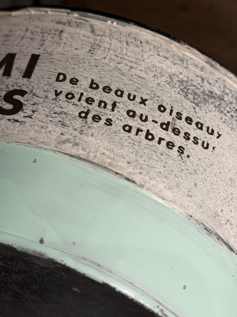 Op de afbeelding: Een cilindrische houder met een textuurrijk, gebroken wit bovengedeelte met de zwarte tekst 'De beaux oiseaux volent au-dessus des arbres.' Het onderste gedeelte is glad, mintgroen van kleur en heeft een donkere rand aan de onderkant.
