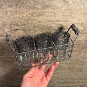 May include: A decorative metal wire basket holding three clear glass tumblers. Each tumbler has a decorative black pattern. The basket has a handle with wooden accents. The basket is held by a person's hand.
