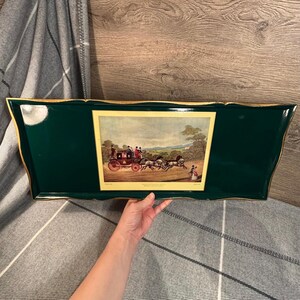 May include: A rectangular, dark green serving tray with a gold trim. The tray features a framed print of a horse-drawn carriage with passengers. The tray is being held by a person.