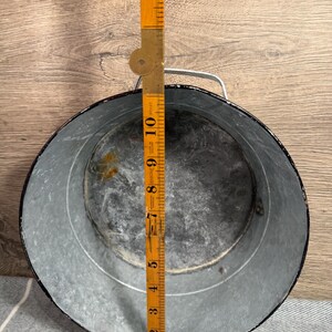 Op de afbeelding: Een ronde gegalvaniseerde metalen bak met een zwarte rand en een zilverkleurig metalen handvat. Een gele opvouwbare houten liniaal is verticaal in het midden geplaatst en toont een diameter van ongeveer 26,5 centimeter voor de opening.